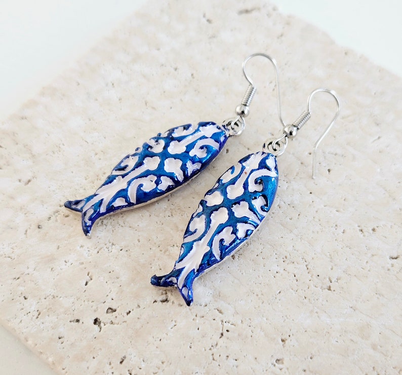 May include: A pair of blue and white fish-shaped earrings with a silver hook. The fish have a white filigree design on a blue background.