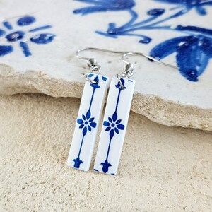 May include: A pair of white ceramic earrings with a blue and white floral pattern. The earrings are dangle style with silver hooks.