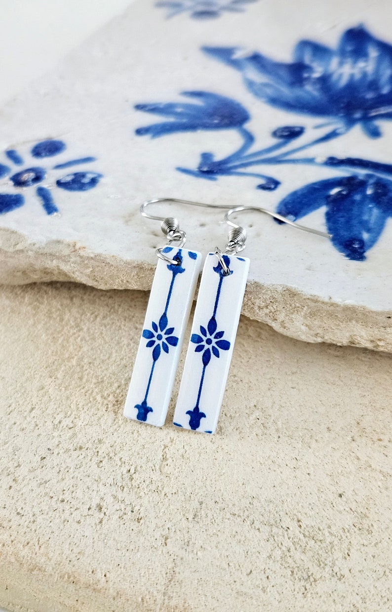 May include: A pair of white ceramic earrings with a blue and white floral pattern. The earrings are dangle style with silver hooks.