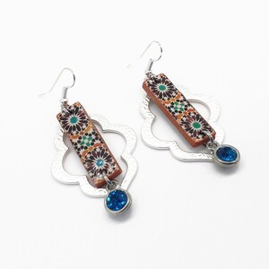 Peut inclure: Une paire de boucles d'oreilles en argent avec un motif g&eacute;om&eacute;trique. Chaque boucle d'oreille pr&eacute;sente un pendentif rectangulaire avec un motif de carrelage marocain color&eacute; et un pendentif en cristal bleu.