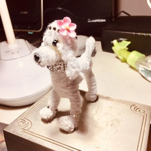 May include: A white fluffy poodle made of pipe cleaners with a pink flower on its head and a silver rhinestone collar. The poodle is standing on a silver box with a decorative design.