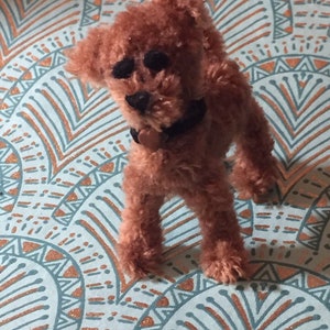 Puede incluir: Un juguete para perro de peluche marrón y peludo, con ojos negros y un collar negro. El juguete está de pie sobre una tela con estampado azul y cobre.