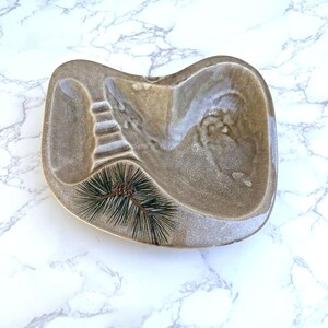 May include: A kidney-shaped ceramic dish with a glossy tan and brown glaze. The surface features a molded ribbed section on the left and a recessed area on the right. A small, dark green pine needle sprig is embedded into the lower left section.