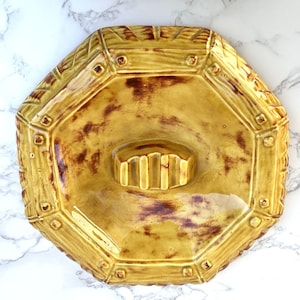 Puede incluir: Un plato de cerámica octogonal vintage en un tono amarillo cálido con acentos marrones. El plato presenta un centro rectangular elevado y un borde decorativo texturizado con vetas de madera simuladas y detalles de remaches. Una pieza única.