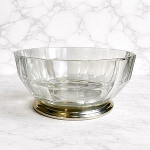 Puede incluir: Un cuenco de vidrio transparente con un diseño geométrico. El cuenco se asienta sobre una base plateada con un borde dorado. El cuenco está vacío y parece estar hecho de vidrio grueso. El fondo es un patrón de mármol blanco.