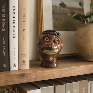 May include: A brown glazed ceramic sculpture of a human head with a glossy finish, featuring prominent round eyes, a distinct dark hair detail on top, and a wide, light green painted mouth, positioned as a decorative object on a wooden shelf.