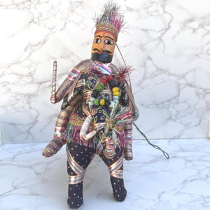 May include: A handcrafted puppet doll with a painted wooden face, adorned with a colorful headdress and elaborate clothing. The doll's attire features a navy blue fabric with gold accents and a beaded necklace. It holds a small, decorative staff.
