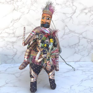 May include: A handcrafted puppet with a painted wooden face, adorned with a crown and mustache. The puppet wears a detailed costume with gold trim, colorful beads, and tassels. It holds a stick and has strings attached for manipulation.