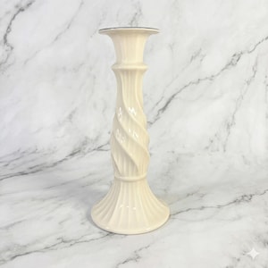 May include: Ivory-colored candlestick holder with a twisted, fluted design. The base and top flare out, and the top has a dark rim. The candlestick is made of ceramic or porcelain and is suitable for a standard-sized candle.