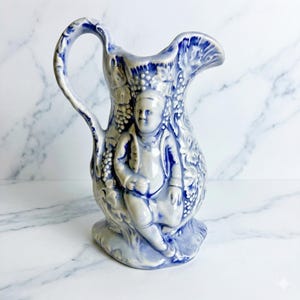 Puede incluir: Jarra de cerámica azul y blanca con un diseño en relieve detallado. La jarra presenta una figura sentada, vides y un asa. La combinación de colores es clásica, adecuada para exhibir o usar.