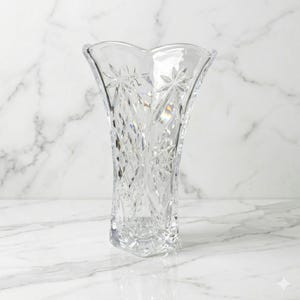 May include: Clear crystal vase with a flared top and starburst and diamond-cut patterns. The vase is approximately 10 inches tall and sits on a white marble surface. The vase is empty and ready to be filled with flowers.