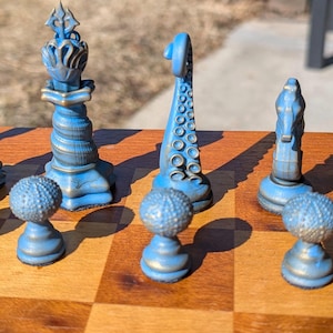 May include: A unique chess set with blue and gold pieces. The pieces have a nautical theme, with tentacle, crown, and spiral designs. The chess board is made of wood and has a classic checkerboard pattern.