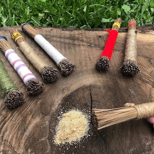 May include: A set of six natural fiber brushes with different colored handles. The brushes are made of natural fibers and are tied together with twine. The brushes are arranged on a wooden surface. There is a pile of sawdust in the lower right corner of the image.