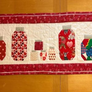 Puede incluir: Un camino de mesa acolchado con diseños de tarros. Los tarros están llenos de patrones de corazones, lunares y otros diseños festivos. El camino de mesa tiene un borde rojo con patrones de corazones y un fondo blanco.