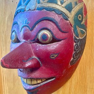 May include: A carved wooden mask with a red face, a long nose, and gold-colored teeth. The mask has blue and gold accents around the eyes and forehead. The mask is likely used for cultural or theatrical purposes.