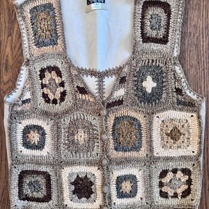 May include: A sleeveless vest constructed from a patchwork of crocheted granny squares in shades of brown, beige, gray, and cream. The garment features a front button closure, a scoop neckline, and a label reading "SABRE HALL" at the inner collar.