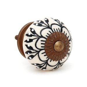 May include: A round ceramic cabinet knob featuring a cream-colored base with black hand-painted floral patterns and white dot accents. The center of the knob has a circular, ridged, antique bronze-toned metal plate with a matching central screw.