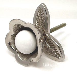 May include: A silver-toned cabinet knob with a flower and leaf design. The knob features a white, spherical center and a threaded post for installation. The leaves have a detailed, textured pattern.