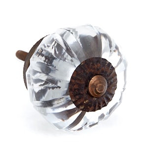 May include: A clear, faceted glass cabinet knob with a rusty brown metal base and screw. The knob has a rounded shape with multiple facets, reflecting light. The metal base has a circular shape with a textured surface.