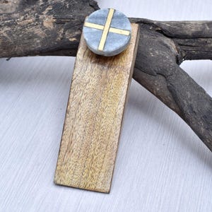 May include: A wooden rectangular drawer pull with a round, gray marble knob. The knob features a gold cross design. The wood has a light brown color with visible grain. The hardware is likely for a cabinet or drawer.
