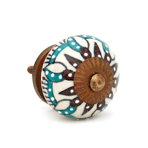 May include: A round ceramic cabinet knob featuring a white base with a hand-painted pattern of dark brown and teal petals accented by small white dots. The center of the knob has a circular, textured, antique brass-colored metal plate.
