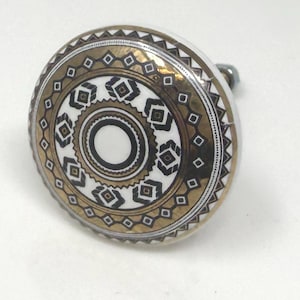 May include: A decorative cabinet knob with a white, gold, and black geometric design. The knob features a circular face with concentric patterns and a silver-toned stem. The design includes diamond and circular shapes.