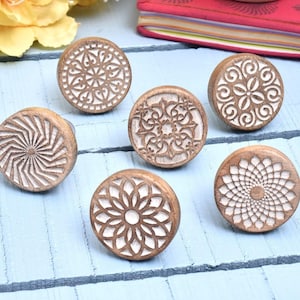 May include: A collection of six wooden cabinet knobs with intricate, carved white designs on a brown background. The knobs feature various patterns, including floral and geometric shapes, and are arranged on a light blue wooden surface.