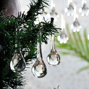 May include: Clear glass teardrop-shaped ornaments hanging from a green Christmas tree. The ornaments reflect the surrounding environment, creating a mirrored effect. Other similar ornaments are visible in the background, suggesting a festive holiday theme.