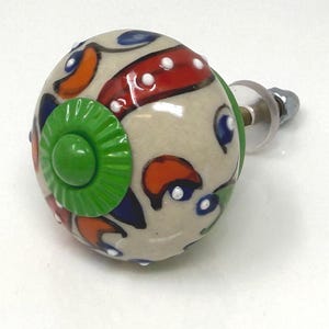 May include: A decorative ceramic cabinet knob with a beige base, featuring a colorful design of red, orange, blue, and green accents. The knob has a round shape with a metal screw for installation. The design is hand-painted.