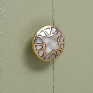 May include: A round brass cabinet knob with a mother-of-pearl inlay. The knob features a central star-shaped design surrounded by triangular segments. The knob is mounted on a light green surface.