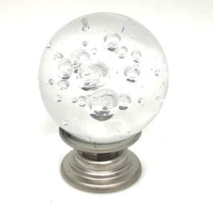 May include: A clear glass sphere with air bubbles, mounted on a brushed metal base. The sphere is round and has a smooth surface. The base is circular with a tiered design. Decorative accent for furniture.