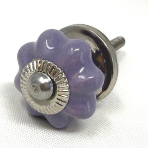 May include: A lavender ceramic cabinet knob with a flower-shaped design. The knob has a silver metal base and a screw for installation. The decorative hardware is suitable for drawers, cabinets, or furniture.