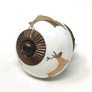 May include: White ceramic cabinet knob with a bronze-colored metal base and deer illustrations. The knob features a bronze-colored center and a decorative bronze-colored ring around the center. The deer are in a gold color.