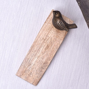 May include: A rectangular wooden serving board with a bronze-colored bird figurine attached to the top. The board has a natural wood grain finish. The bird is detailed with etched feathers and a small beak. The board is designed for serving appetizers or small dishes.