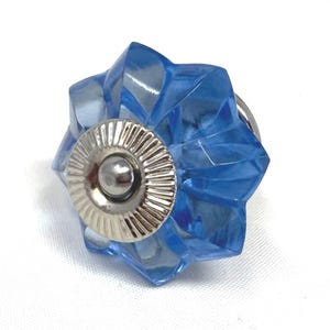 May include: A blue, star-shaped cabinet knob with a silver-colored base and a small, round center. The knob is made of a transparent material, allowing light to pass through. The design is faceted, giving it a sparkling appearance.