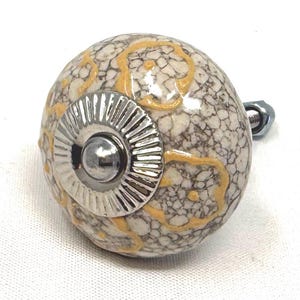 May include: A decorative cabinet knob with a round, speckled, off-white and brown ceramic body. The knob features a silver metal base and a silver center. Yellow swirls accent the ceramic. The knob is attached to a bolt.