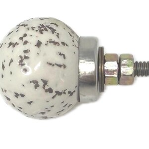 May include: A round cabinet knob featuring a glossy white ceramic surface with a speckled dark brown pattern. The knob is attached to a silver-toned metal base with a threaded bolt, two metal washers, and two hexagonal nuts for secure installation.