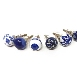 May include: A collection of eight ceramic cabinet knobs. The knobs are primarily white and navy blue, featuring various patterns such as floral designs, stripes, and polka dots. Each knob has a metal screw for easy installation.