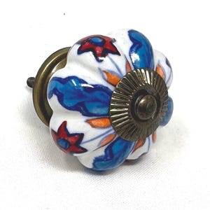 May include: Decorative ceramic cabinet knob with a floral design. The knob features a white base with blue, red, and orange painted details. It has a brass-colored metal base and screw.