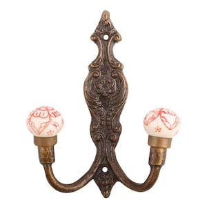 May include: Ornate bronze-colored wall hook with two curved arms. Each arm ends with a small, round, white ceramic knob decorated with a red floral pattern. The hook has a decorative, vertical backplate.