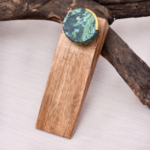 May include: A wooden doorstop with a round, marbled stone top in shades of green and blue, accented with a gold-colored band. The doorstop is made of light brown wood.