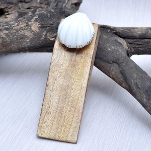 May include: A wooden doorstop with a white seashell accent. The doorstop is rectangular and made of light brown wood. The seashell is white with subtle brown detailing. The doorstop is designed to hold a door open.