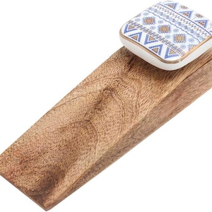 May include: A wooden doorstop with a decorative ceramic top. The ceramic top is square with a white background and blue and gold geometric patterns. The doorstop is made of light brown wood.