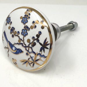 May include: White ceramic cabinet knob with a gold rim and a decorative design featuring blue flowers, a blue bird, and gold branches. The knob has a metal screw for installation.