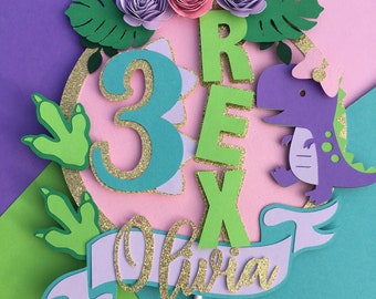 Può includere: Un topper per torta a tema dinosauro rosa e verde con il numero 3, la parola "Rex" e il nome "Olivia" in glitter dorato. Il topper presenta un dinosauro viola con un fiocco rosa, foglie verdi e fiori rosa.