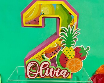 May include: A pink and yellow number 2 birthday decoration with a fruit theme. The number is filled with images of bananas, watermelon, and other fruits. The name "Olivia" is written in cursive. A pineapple, strawberry, and orange are attached to the number.