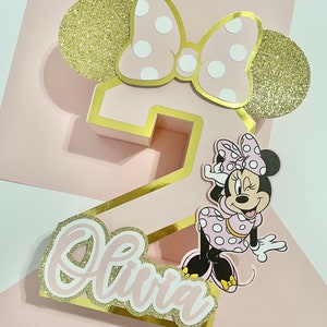 K&ouml;nnte beinhalten: Rosa-goldener Zahl-Zwei-Kuchentopper mit einer Minnie Mouse Figur, einer rosa Schleife mit wei&szlig;en Punkten und dem Namen Olivia in goldenen Glitzerbuchstaben.