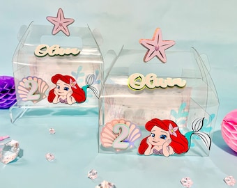 May include: Two clear plastic favor boxes decorated with a mermaid theme. Each box has a pink starfish on top, a mermaid character, a seashell with the number 2, and the name "Olivia" written in green and yellow letters.