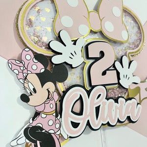 K&ouml;nnte beinhalten: Rosa-goldener Kuchentopper im Minnie Mouse-Design mit einer rosa Schleife, wei&szlig;en Punkten und der Zahl 2. Der Topper enth&auml;lt auch den Namen Olivia in rosa Schrift.