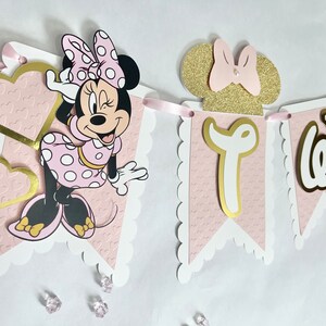 K&ouml;nnte beinhalten: Rosa-goldener Minnie Mouse-Banner mit dem Wort "Zwei" in goldenen Buchstaben. Der Banner hat einen rosa Hintergrund mit wei&szlig;en gewellten R&auml;ndern und eine Minnie Mouse-Figur.